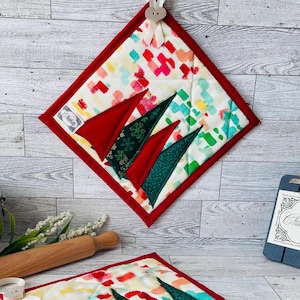 May include: A quilted Christmas-themed potholder with a red border and a colorful abstract pattern. The potholder features appliqued red and green Christmas tree shapes. A small tag is attached to the potholder.