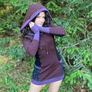 Puede incluir: Una mujer con un vestido con capucha morado y marrón con un patrón geométrico en blanco y negro. Está de pie en un entorno boscoso.