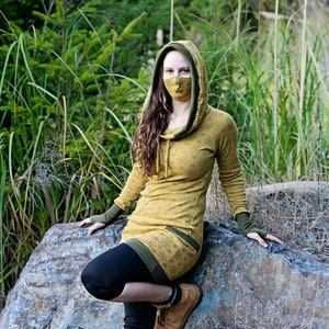 Puede incluir: Una persona lleva un vestido amarillo con capucha, ribete verde y diseño estampado. Lleva una mascarilla facial y está sentada en una gran roca en una zona cubierta de hierba.