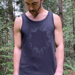 May include: Black tank top with a geometric design on the front. The tank top is paired with black sweatpants. The person is standing in a forest setting with tall trees and green foliage.