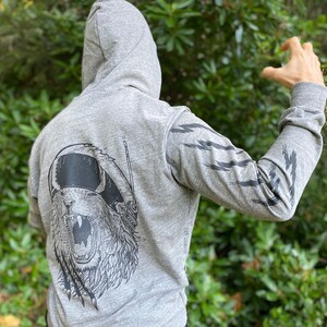 Puede incluir: Sudadera con capucha gris con un gráfico de un oso rugiente con auriculares en la espalda. Las mangas tienen diseños de rayos negros. La persona lleva pantalones negros.
