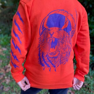 Puede incluir: Sudadera con capucha naranja con un diseño de tigre morado en la espalda y marcas de garras moradas en la manga. La sudadera está hecha de un material suave y tiene una capucha con cordón.