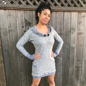 Puede incluir: Un vestido gris con capucha y estampado geométrico. El vestido tiene cuello de pico y mangas largas con un detalle de botón en la parte delantera. El vestido es entallado y tiene una longitud mini.