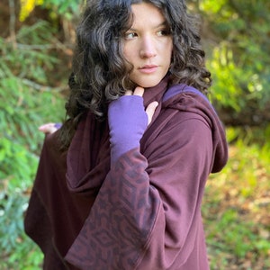 Puede incluir: Un poncho marrón con capucha, con un patrón geométrico en las mangas y un guante sin dedos morado. El poncho está hecho de una tela suave y cómoda y tiene un corte holgado.