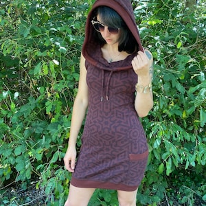 Puede incluir: Un vestido marrón con capucha y un patrón geométrico. El vestido tiene una capucha con cordón y un bolsillo en el lateral.