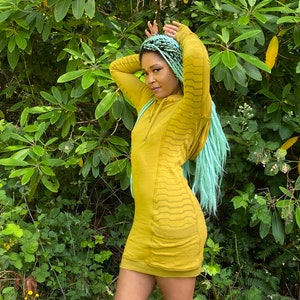 Puede incluir: Una mujer con un vestido amarillo mostaza con capucha y un patrón geométrico. Tiene el pelo azul y verde y está de pie frente a un fondo verde de hojas.