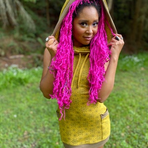 Puede incluir: Minivestido con capucha verde oliva con un estampado geométrico. El vestido tiene una capucha con cordón y dos bolsillos delanteros. El ribete y el forro de la capucha son marrones. La modelo tiene el pelo rosa brillante.