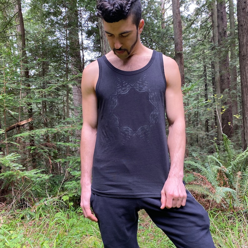 May include: Black tank top with a geometric design on the front. The tank top is paired with black sweatpants. The person is standing in a forest setting with tall trees and green foliage.