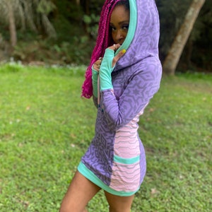 Puede incluir: Un vestido morado y rosa con capucha y un patrón geométrico. El vestido tiene mangas largas y una silueta ajustada. La modelo lleva el vestido con trenzas rosas y botas marrones.