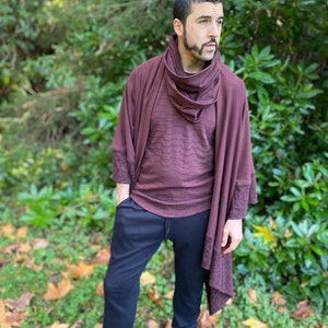 Puede incluir: Una persona con un chal burdeos y una camisa de manga larga con un patrón sutil, combinados con pantalones negros y botas. El conjunto se presenta sobre un fondo de follaje verde y hojas caídas.