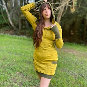 Puede incluir: Un vestido amarillo y verde con capucha y un patrón ondulado. El vestido tiene mangas largas y un bolsillo canguro. La modelo lleva el vestido en un entorno de bosque.