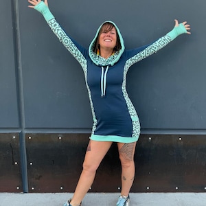 Puede incluir: Una mujer lleva una sudadera con capucha azul turquesa y verde menta con un patrón geométrico. La sudadera tiene cuello de pico y mangas largas. La mujer está de pie frente a una pared gris.