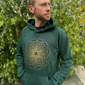 May include: A dark green hoodie with a gold geometric design on the front. The hoodie has a kangaroo pocket and a drawstring hood. The design is a detailed, symmetrical pattern with a star in the center. The person is wearing a black shirt underneath.
