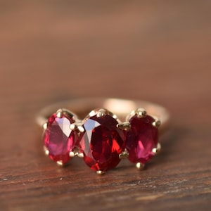 May include: A gold ring featuring three oval-cut, deep red gemstones. The gemstones are set closely together, creating a cluster design. The ring is displayed on a wooden surface.