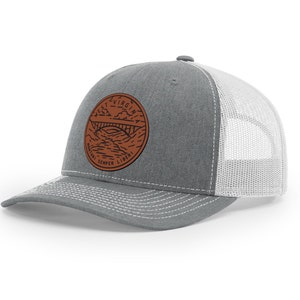 May include: A gray trucker hat with a white mesh back and a curved brim. The front features a circular brown patch with a bridge design and the words "West Virginia" and "Montani Semper Liberi" in a circular pattern.