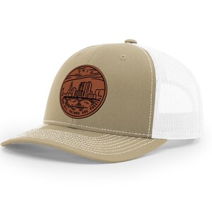 May include: A tan and white trucker hat with a brown leather patch featuring a skyline and the text "OHIO WITH GOD ALL THINGS ARE POSSIBLE".