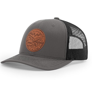 May include: A gray and black trucker hat with a brown leather patch featuring a mountain scene and the text "Alaska, Where the Future is Wild".