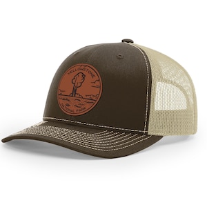 May include: A brown and beige trucker hat with a brown leather patch featuring the Yellowstone National Park logo. The logo depicts a geyser erupting with a tree in the background.