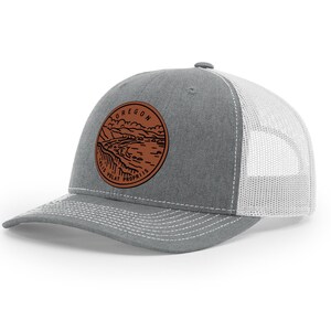 May include: A gray and white trucker hat with a brown leather patch featuring a landscape scene and the text "OREGON COAST VOLCANIC PROVINCE".