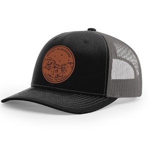 May include: Black and gray trucker hat with a brown leather patch featuring a silhouette of Mount Rushmore National Memorial with the text "Mount Rushmore National Memorial".