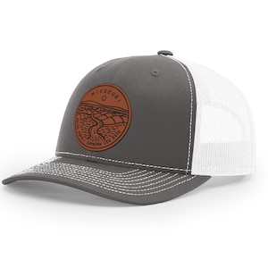 May include: A gray and white trucker hat with a brown leather patch featuring a state outline and the text "MISSOURI O SALUS POPULI SUPREMA LEX ESTO".