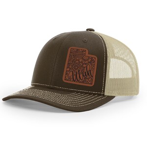 May include: A brown and tan trucker hat with a brown leather patch featuring a detailed outline of the state of Utah with the word "Utah" written in cursive.
