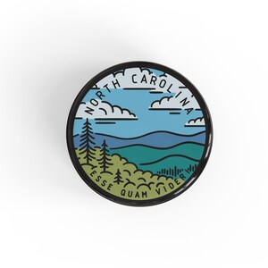 Pode incluir: Um emblema redondo com borda preta, apresentando um design de paisagem com o texto "NORTH CAROLINA" e "ESSE QUAM VIDERI". O design inclui um c&eacute;u azul, nuvens, colinas verdes e pinheiros.