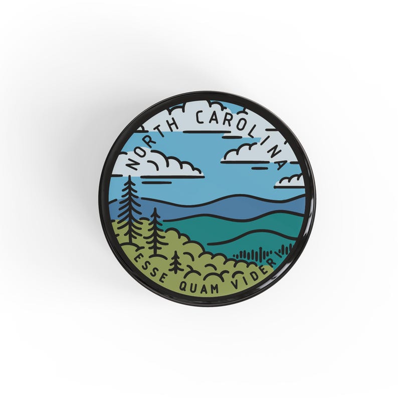 Pode incluir: Um emblema redondo com borda preta, apresentando um design de paisagem com o texto "NORTH CAROLINA" e "ESSE QUAM VIDERI". O design inclui um c&eacute;u azul, nuvens, colinas verdes e pinheiros.