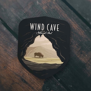 May include: A black, shield-shaped sticker featuring a stylized illustration of Wind Cave National Park. The design includes a cave entrance, a bison, and the text "WIND CAVE National Park" in white lettering. The sticker is set against a dark wooden surface.
