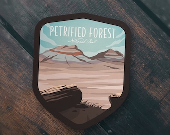 May include: A brown and tan illustrated sticker with a shield shape. The sticker features a landscape scene of a desert with a blue sky and white clouds. The text "Petrified Forest National Park" is at the top of the sticker.
