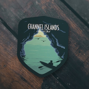 May include: A dark blue and green sticker with a silhouette of a person kayaking through a cave opening. The sticker says "Channel Islands National Park" at the top.