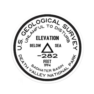 May include: A black and white round sticker with the text "U.S. GEOLOGICAL SURVEY UNLAWFUL TO DISTURB" around the edge. In the center is a triangle with the text "ELEVATION BELOW SEA -282 FEET 1994" and "DEATH VALLEY NATIONAL PARK BADWATER BASIN".
