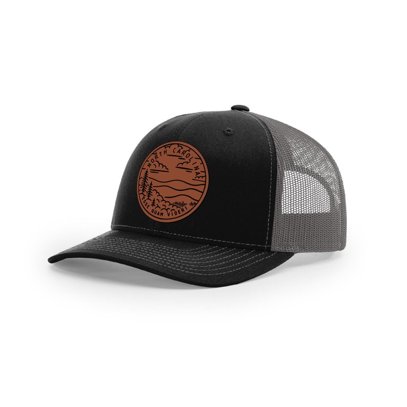 May include: A black and gray trucker hat with a brown leather patch that says "North Carolina" with a mountain scene and the text "The Old North State" underneath.