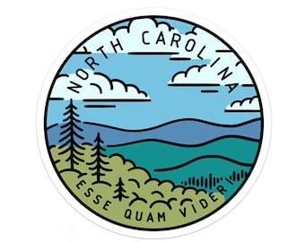 Op de afbeelding: Een ronde sticker met een blauwe lucht, witte wolken, groene bomen en bergen. De tekst "NORTH CAROLINA" staat in een cirkel rond de bovenkant van de sticker. De tekst "ESSE QUAM VIDERI" staat in een cirkel rond de onderkant van de sticker.