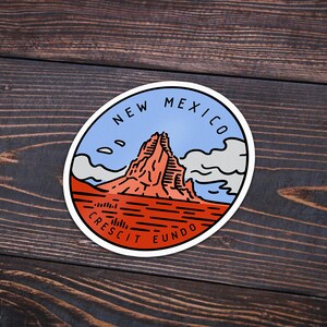 May include: A round sticker with a blue, white, and red design. It features a mountain range with the text "NEW MEXICO" and "CRESCIT EUNDO" around the edge.