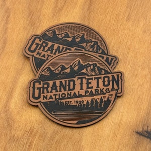May include: Two brown leather patches with the text "GRAND TETON NATIONAL PARK" and an illustration of mountains and trees. The patches are circular with a black border and black text. The text "EST. 1929" is also included.