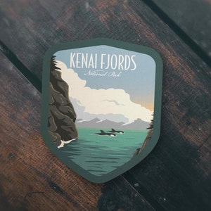 May include: A green and blue illustrated sticker with the text "Kenai Fjords National Park". The sticker features a scenic view of a fjord with mountains in the background and orcas swimming in the water.