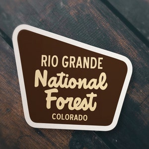 May include: A brown and white sticker with the text "Rio Grande National Forest Colorado" in a retro font.