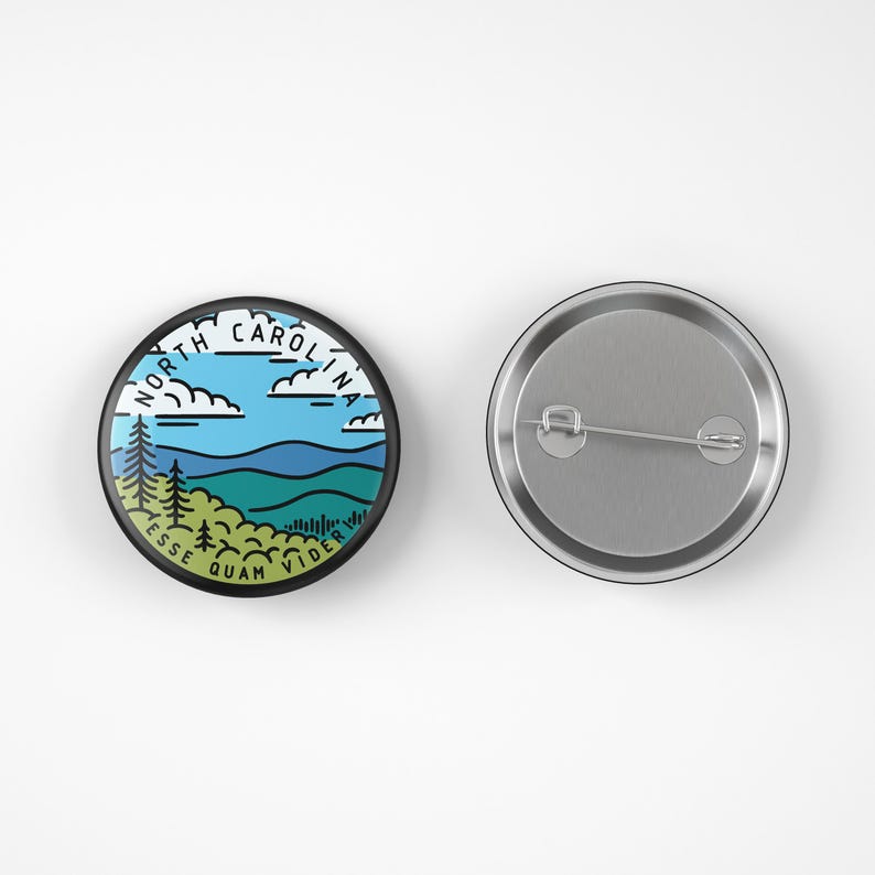 Pode incluir: Um bot&atilde;o redondo com uma borda preta com um design de paisagem. O design inclui c&eacute;us azuis, nuvens brancas, &aacute;rvores verdes e &aacute;gua azul. As palavras "NORTH CAROLINA" e uma frase em latim tamb&eacute;m est&atilde;o inclu&iacute;das.