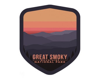 May include: A shield-shaped sticker with a sunset over a mountain range. The text "Great Smoky Mountains National Park" is printed in white on a dark brown background.