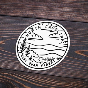 May include: Black and white sticker with a circular design featuring a mountain range, clouds, and trees. The text "NORTH CAROLINA" is written in a circular arc at the top of the sticker. The text "ESSE QUAM VIDERI" is written in a circular arc at the bottom of the sticker.