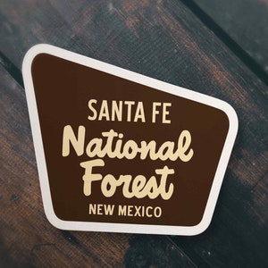 May include: A brown and white sticker with the text "Santa Fe National Forest" and "New Mexico" in a retro style font.