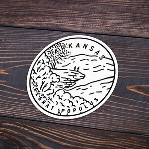 May include: Black and white illustration of a landscape with a river, trees, and hills. The text "KANSAS AD ASTRA PER ASPERA" is written around the edge of the circle.