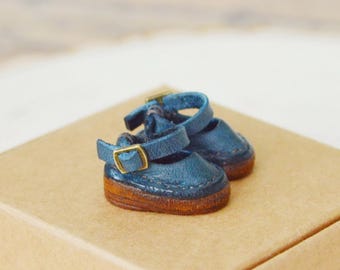 May include: Miniature blue Mary Jane shoes with gold buckles and brown soles rest on a small cardboard box. The shoes are a glossy blue and appear to be made of leather. The box is a light brown color.
