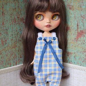 May include: A doll wearing a blue and yellow plaid overall dress with a blue bow. The doll has long brown hair and blue eyes.