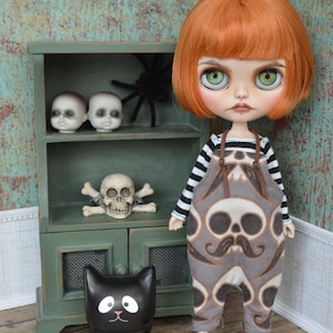 May include: A doll with orange hair and green eyes wearing a black and white striped shirt and gray overalls with a skull and mustache pattern. A black cat figurine and a green cabinet with skulls and a spider are also in the image. The text "Drop Dead Fashions dropdeadfashions.etsy.com" is visible.