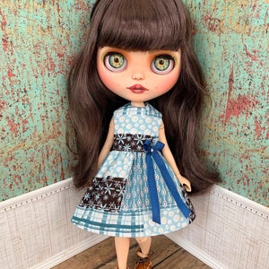 May include: A doll wearing a blue and brown patchwork dress with a blue ribbon. The doll has long brown hair and green eyes. The doll is standing on a wooden floor.