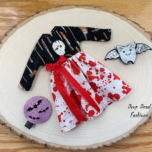May include: A Halloween-themed doll dress with a black and green top featuring a skull and knives, and a white and red skirt with blood splatters. The dress is accompanied by two bat-shaped hair clips, one purple and one silver.