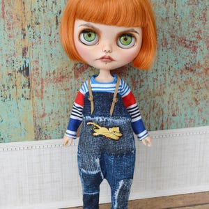May include: A doll with short, bright orange hair wearing a blue and red striped long-sleeved shirt and blue denim overalls with a cat design. The doll is wearing black shoes with buckles. The doll has green eyes and a neutral expression. The background is a turquoise wall with a white trim.