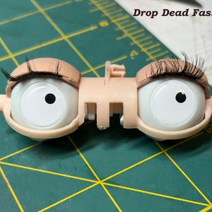 May include: A pair of doll eyes with attached eyelashes. The eyes are white with black pupils and are set in a light tan plastic frame. The text "Drop Dead Fashions" is visible in the upper right corner.
